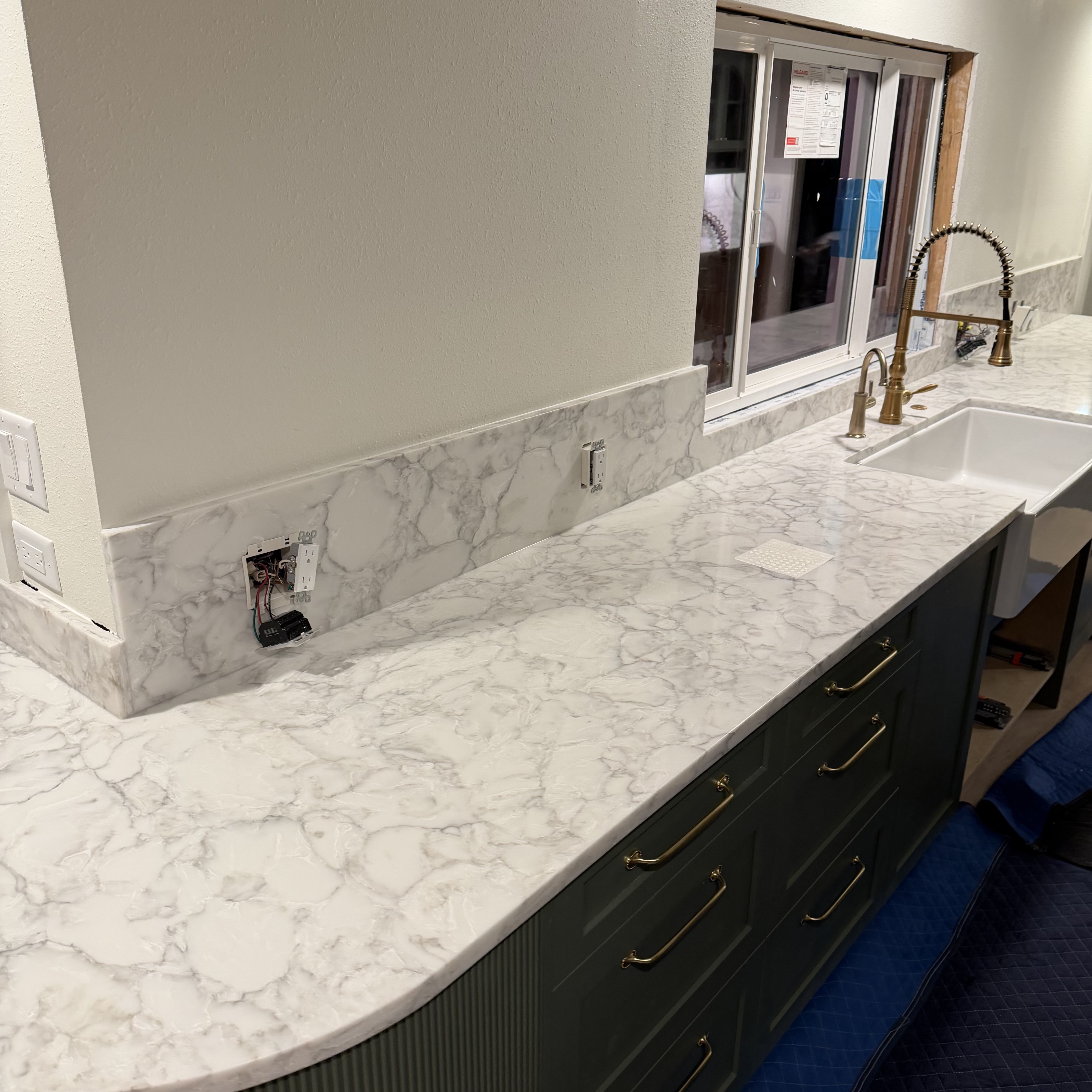 White and gray patterned stone countertop and backsplash with a farmhouse sink and brass faucet