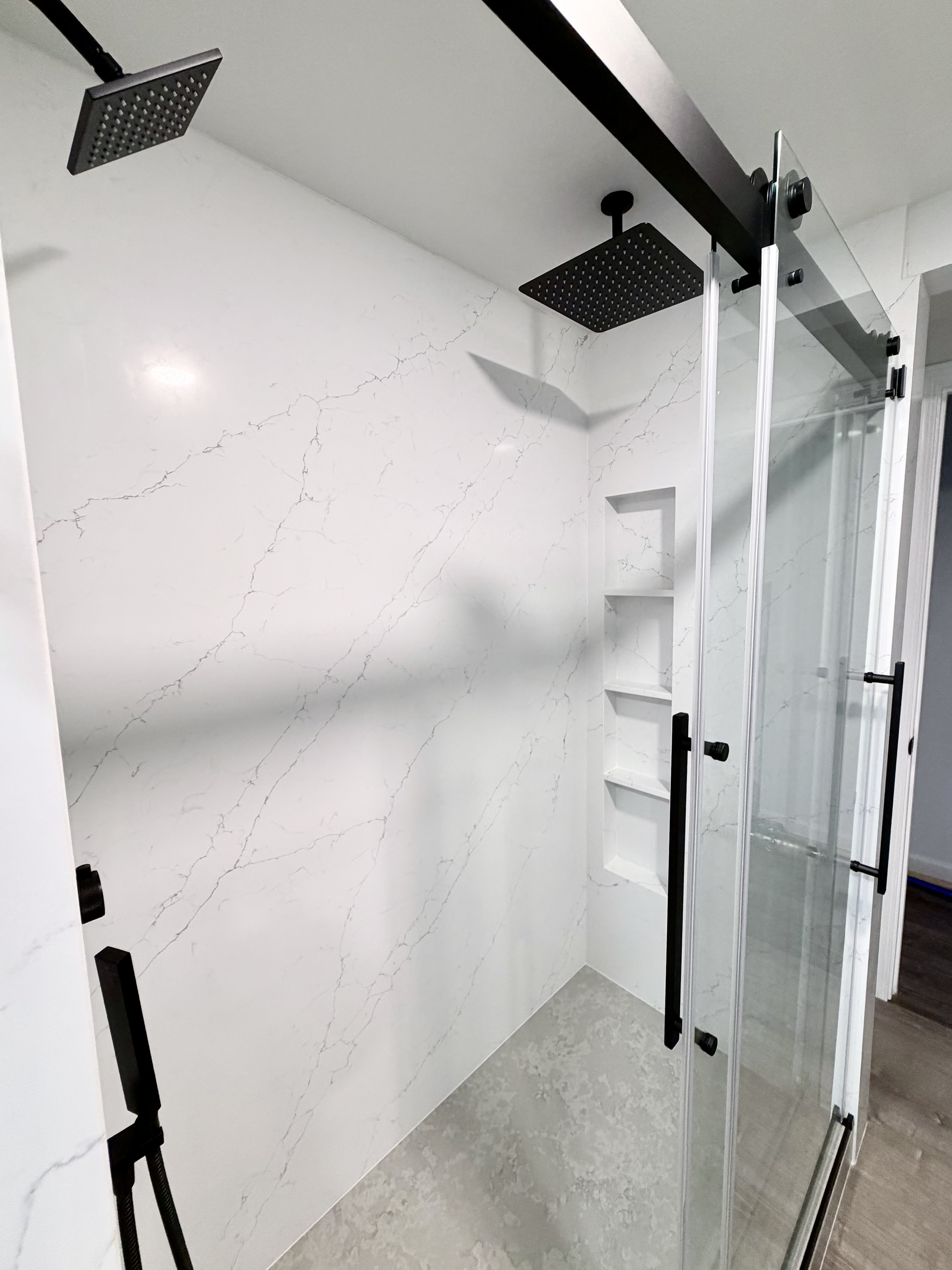 Full-height white veined shower wall surfaces with recessed shelves, black fixtures, and a glass enclosure